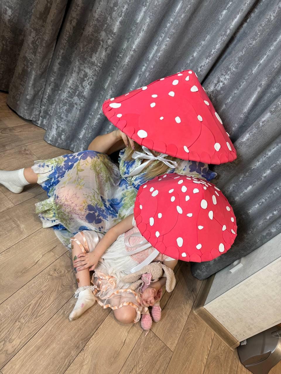 May include: Two red mushroom-shaped hats with white spots. One hat is worn by a person wearing a floral dress, and the other is worn by a child in a white dress. The scene is set indoors with a wooden floor and gray curtains.