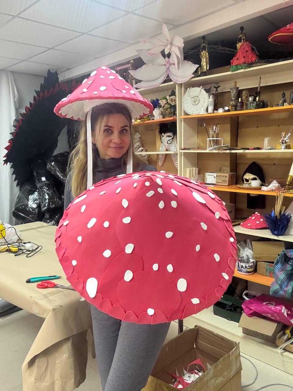 May include: A whimsical mushroom costume with a large, red, rounded cap and a smaller, matching hat. The costume features white polka dots and a white underside. The person is wearing gray leggings and a gray turtleneck.