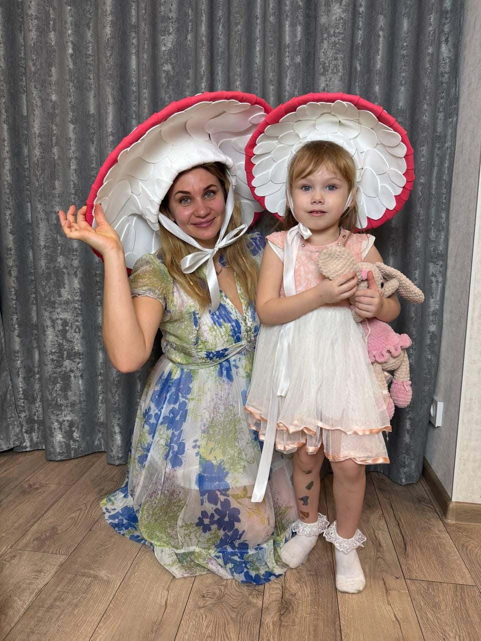 May include: A woman and a child wearing mushroom-shaped hats with white petals and red trim. The woman wears a floral dress, and the child wears a pink and white dress and holds a stuffed bunny. The hats are the focal point of the image.