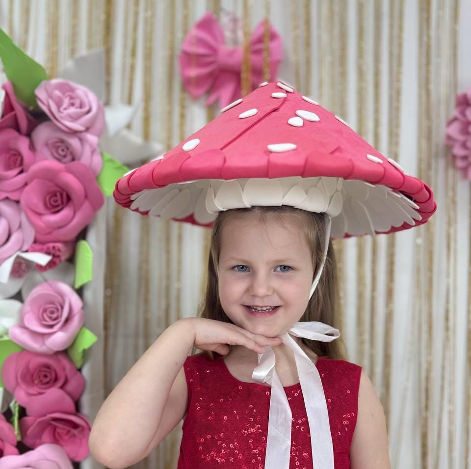 May include: A whimsical mushroom hat with a bright red cap and white polka dots. The hat has a white underside and is secured with a white ribbon. The hat is part of a costume or accessory.
