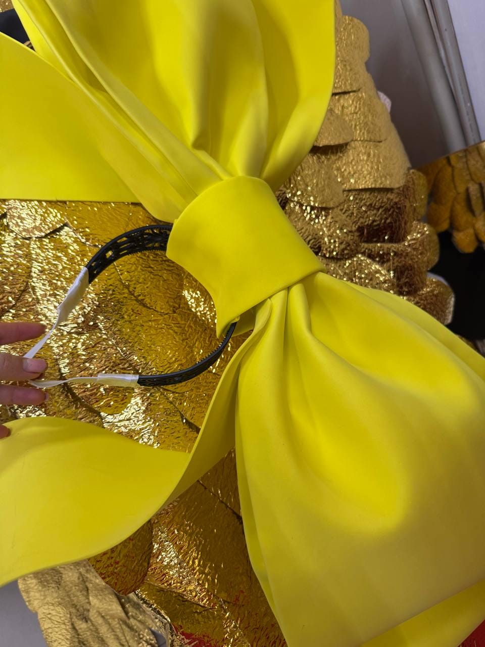 May include: A close-up of a large, bright yellow bow. The bow is made of a soft, pliable material and is tied in a large knot. The background features gold-colored, textured material.