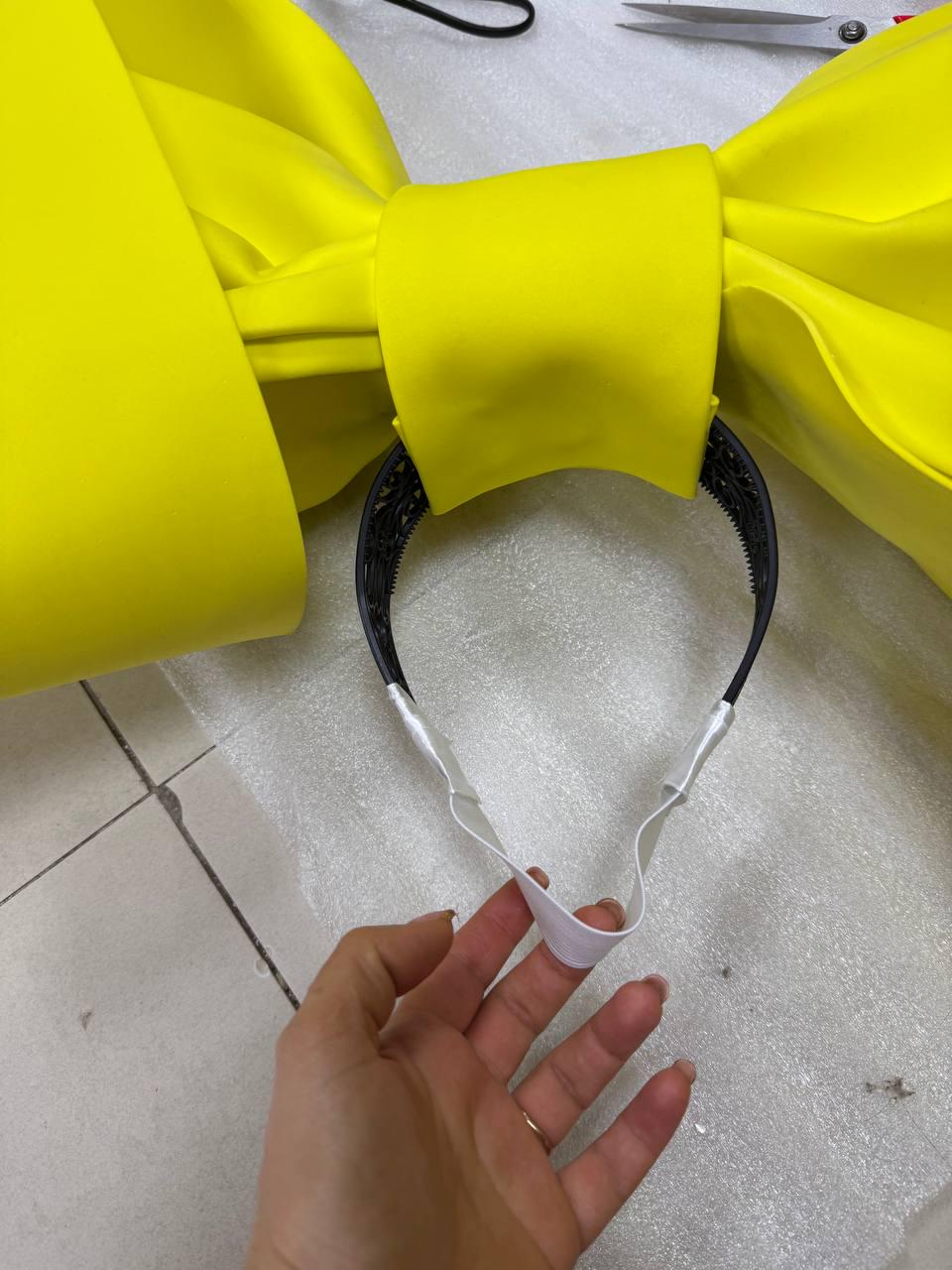 May include: A large, bright yellow bow headband. The bow is made of a smooth, satin-like material. The headband is black with white elastic straps attached to the bow.