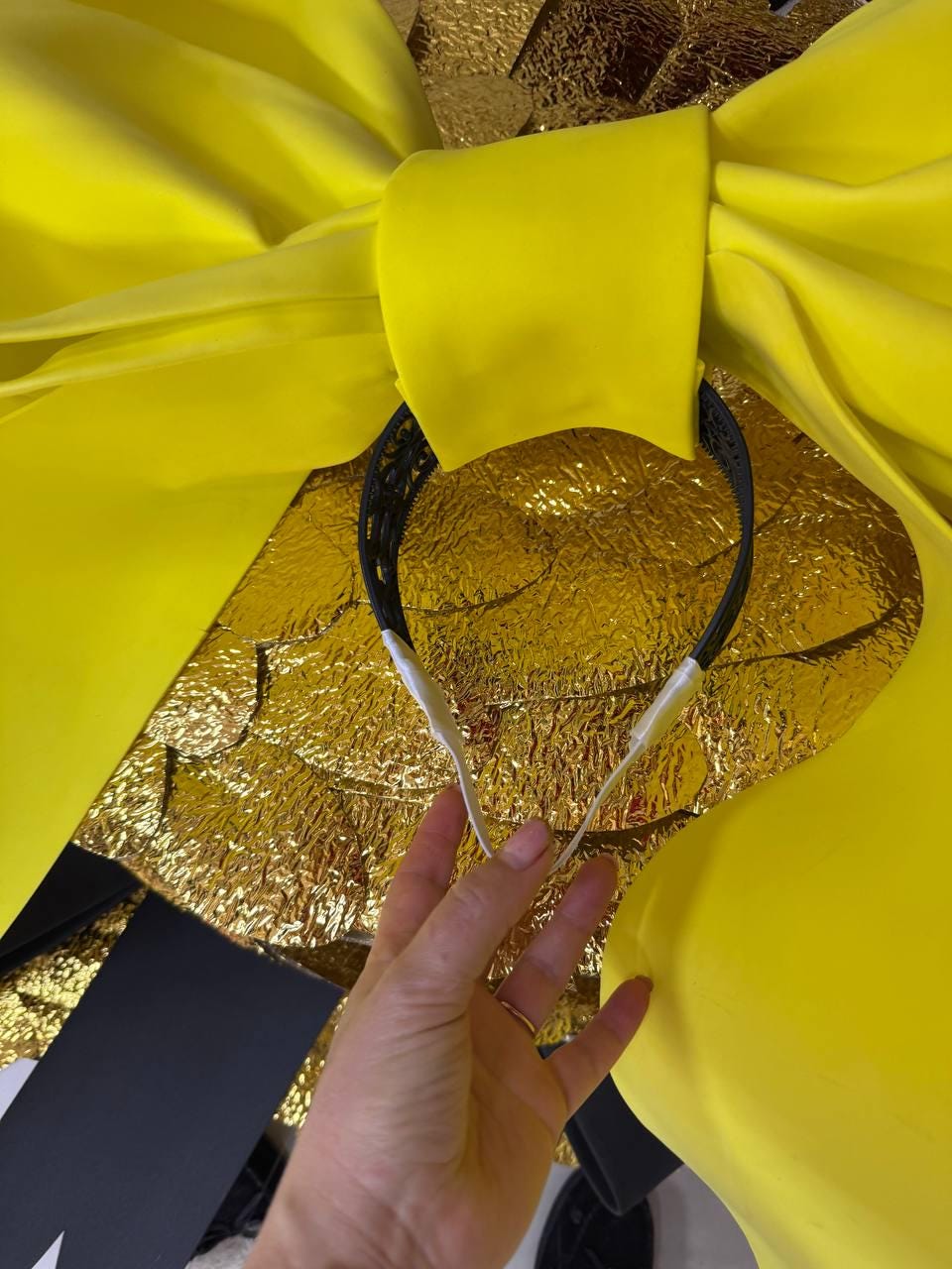 May include: A yellow satin bow with a black headband. The bow is large and has a shiny texture. The headband is black with white accents. The background is gold and black.