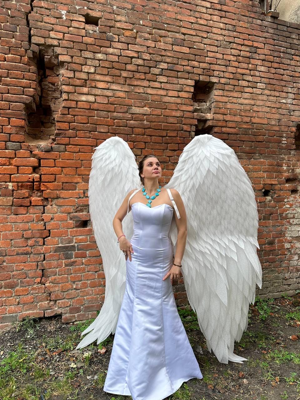 May include: A person wearing a white dress and large white angel wings, standing in front of a brick wall. The person is wearing a turquoise necklace.