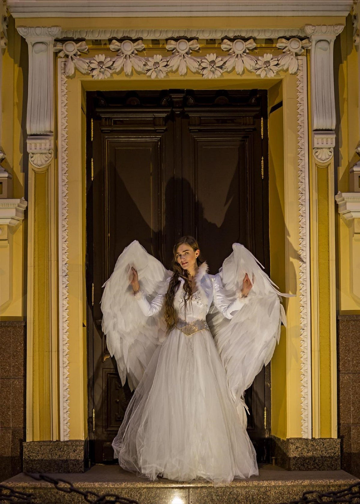 May include: A woman wearing a white dress and angel wings stands in front of a doorway with ornate trim. The doorway is framed by a yellow wall with decorative molding.