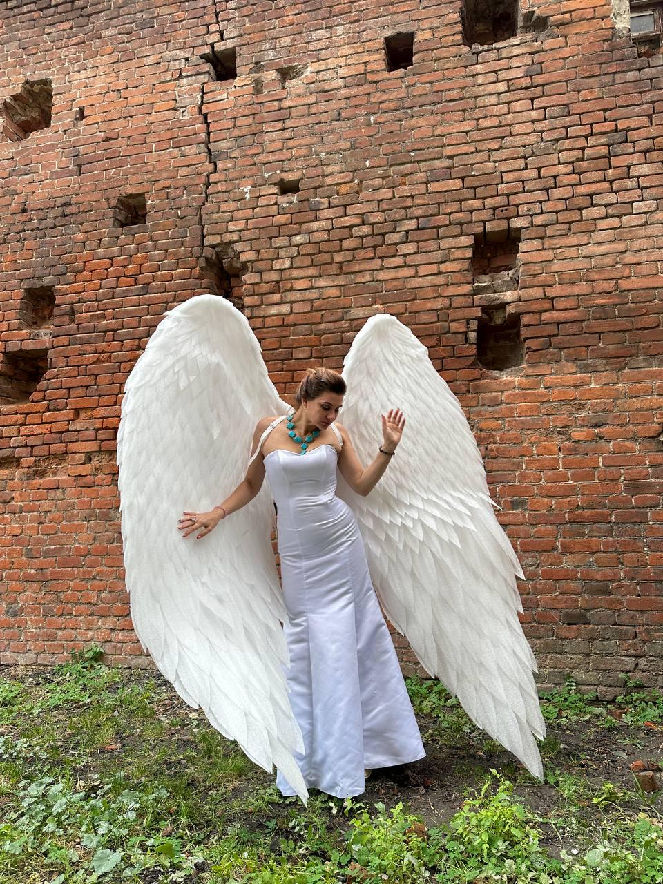 May include: A person wearing a white dress and large white angel wings, standing in front of a brick wall.
