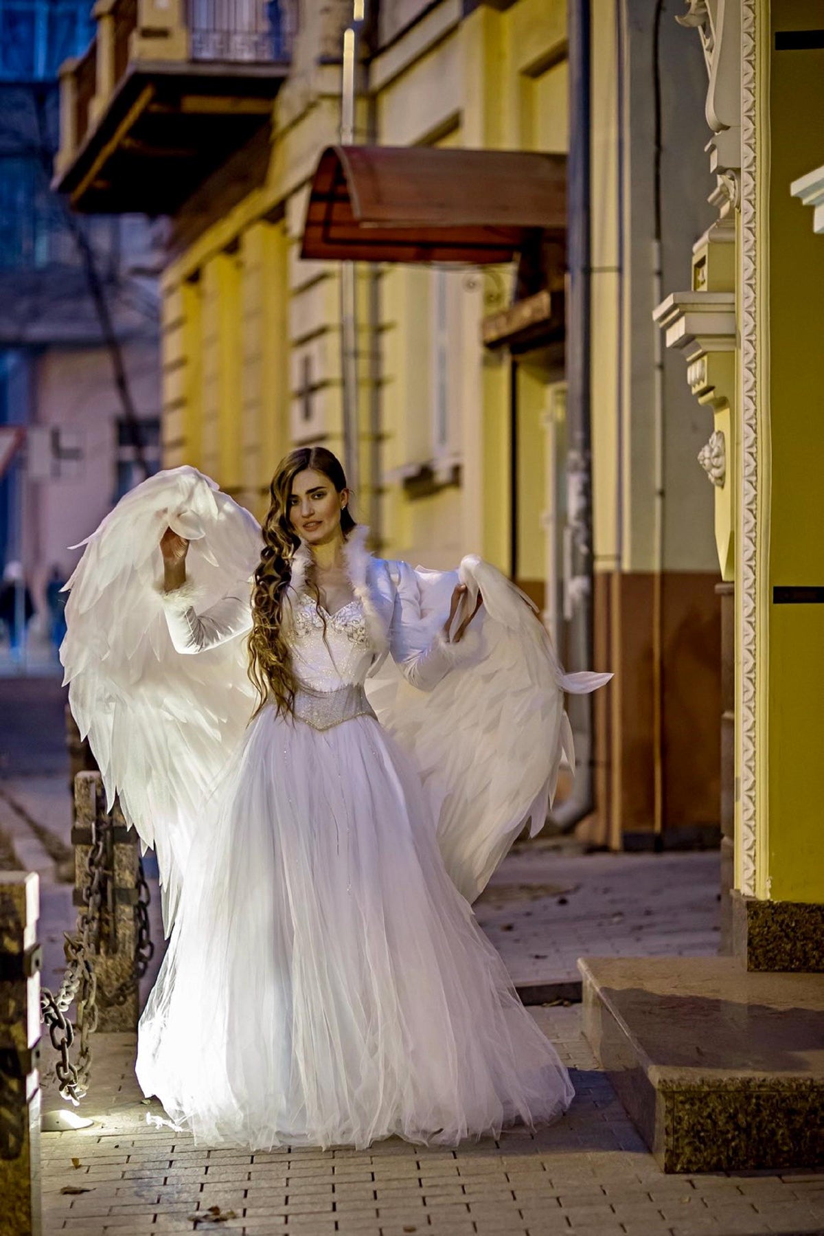 May include: A woman wearing a white dress and angel wings stands in front of a building. The dress is long and flowing, and the wings are large and feathery. The woman is smiling and looking at the camera.