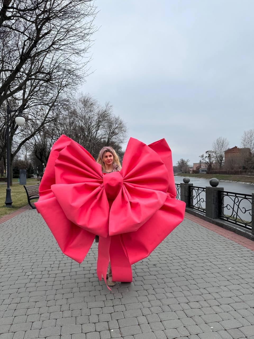 May include: A large, vibrant pink bow, possibly a costume or prop, dominates the image. The bow is the focal point, with a person partially visible behind it. The setting appears to be outdoors, with a paved walkway and trees in the background.