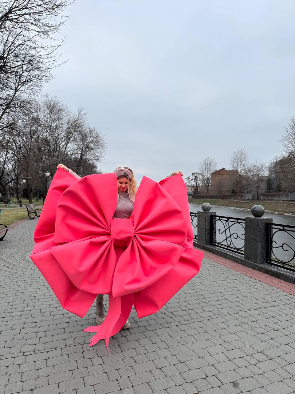 May include: A large, vibrant pink bow, held open against a cloudy sky. The bow is made of a smooth, possibly fabric material, with a detailed, layered design. The bow's size is substantial, creating a striking visual impact.