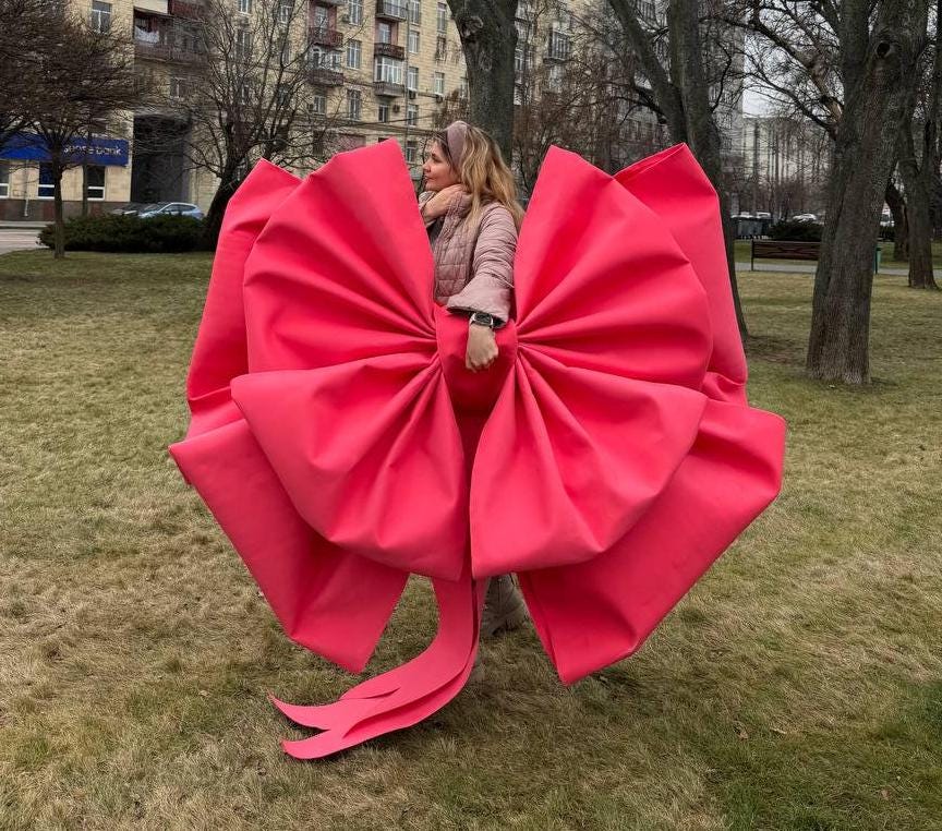 May include: A large, vibrant red bow made of fabric. The bow is oversized and has multiple layers, with long, flowing tails. The bow is the focal point, set against a backdrop of a park with trees and grass.