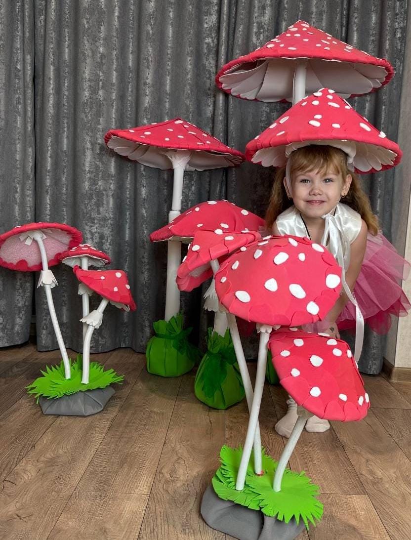 May include: A young girl wearing a white dress and a red and white mushroom hat stands in front of a group of large red and white mushroom decorations. The mushrooms have white stems and green bases.