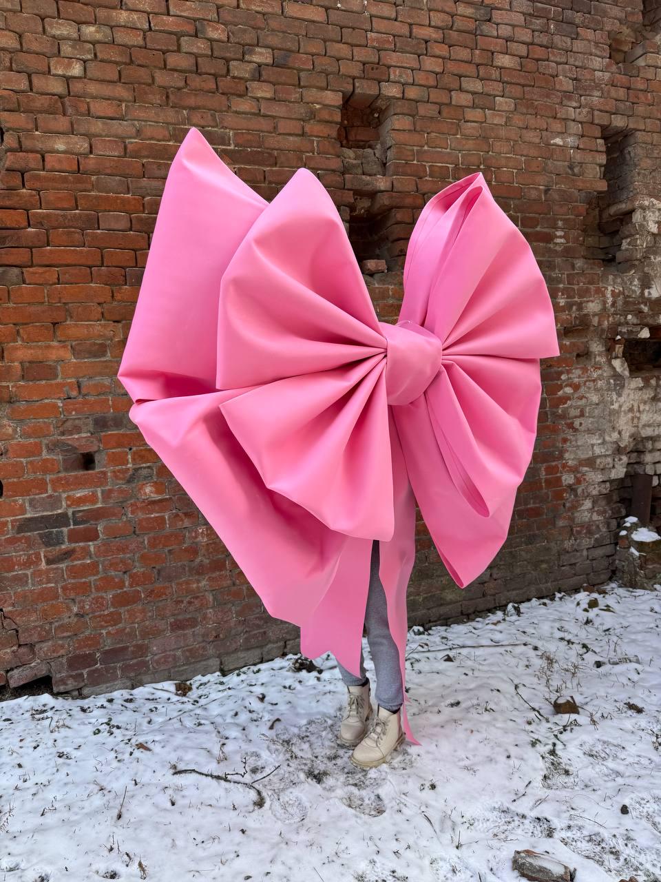 May include: A large pink bow made of fabric is positioned in front of a brick wall. The bow is tied in a knot and the ends of the bow are trailing on the ground. The bow is in front of a person wearing white shoes and gray pants.
