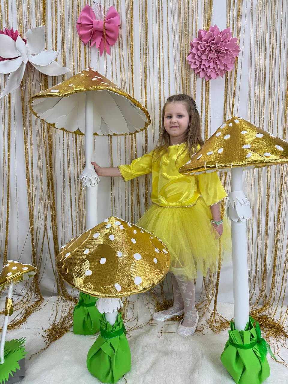 May include: A young girl wearing a yellow dress and white shoes stands in front of a backdrop of gold fringe and pink flowers. She is holding a large, white and gold mushroom prop with white polka dots. There are other mushroom props in the background.