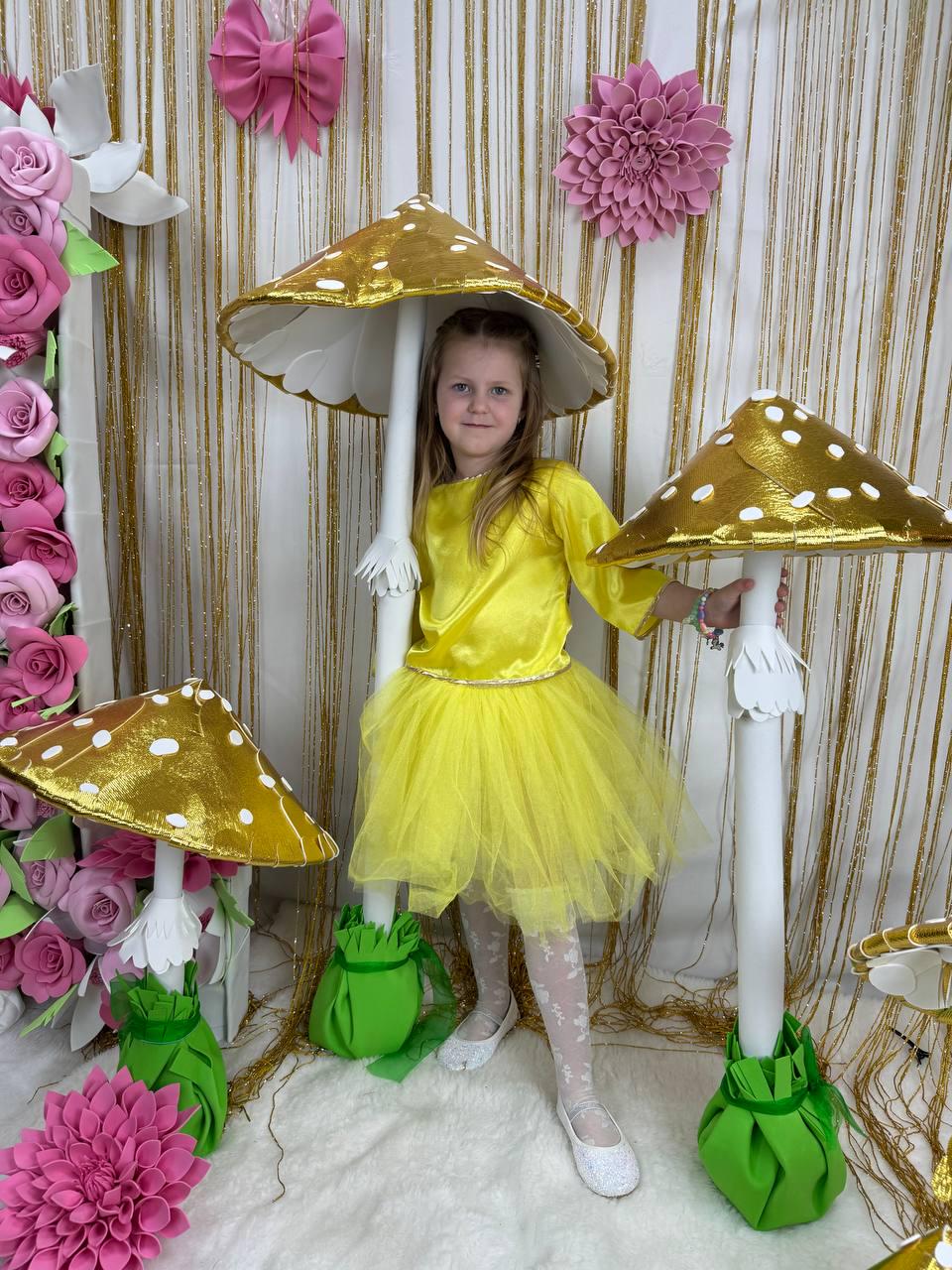 May include: A young girl wearing a yellow dress and white tights stands in front of a backdrop of pink flowers and gold fringe. She is holding a large, gold and white mushroom prop. There are two smaller mushrooms on either side of her.
