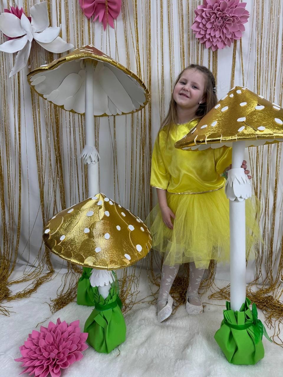 May include: Two large, decorative mushroom props with white and gold accents. The larger mushroom has a white top and a gold base. The smaller mushroom has a gold top with white polka dots and a green base. A young girl in a yellow dress stands between the mushrooms.