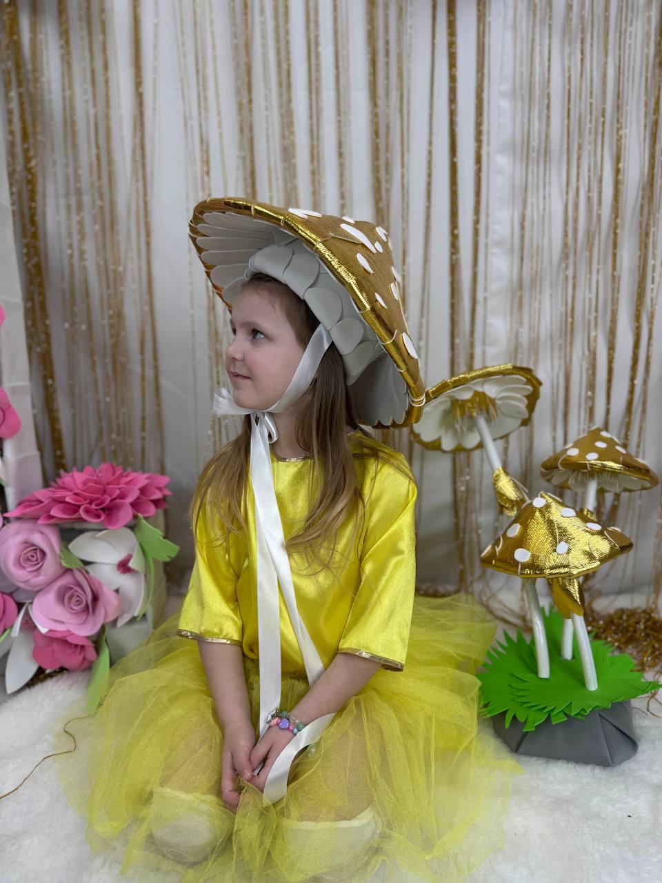 May include: A young girl wearing a yellow dress and a gold mushroom hat with white polka dots. She is sitting on a white surface with a green and gold mushroom prop beside her.