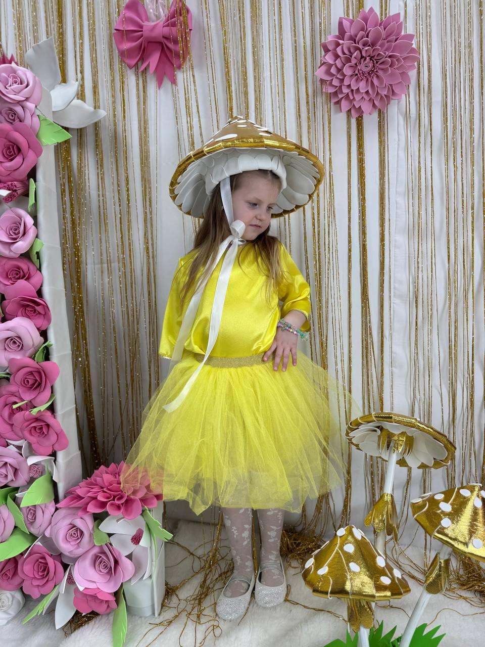 May include: A young girl wearing a yellow tulle skirt and a white and gold mushroom hat. The hat has a white brim and gold accents. The girl is standing in front of a backdrop of pink flowers and gold streamers.
