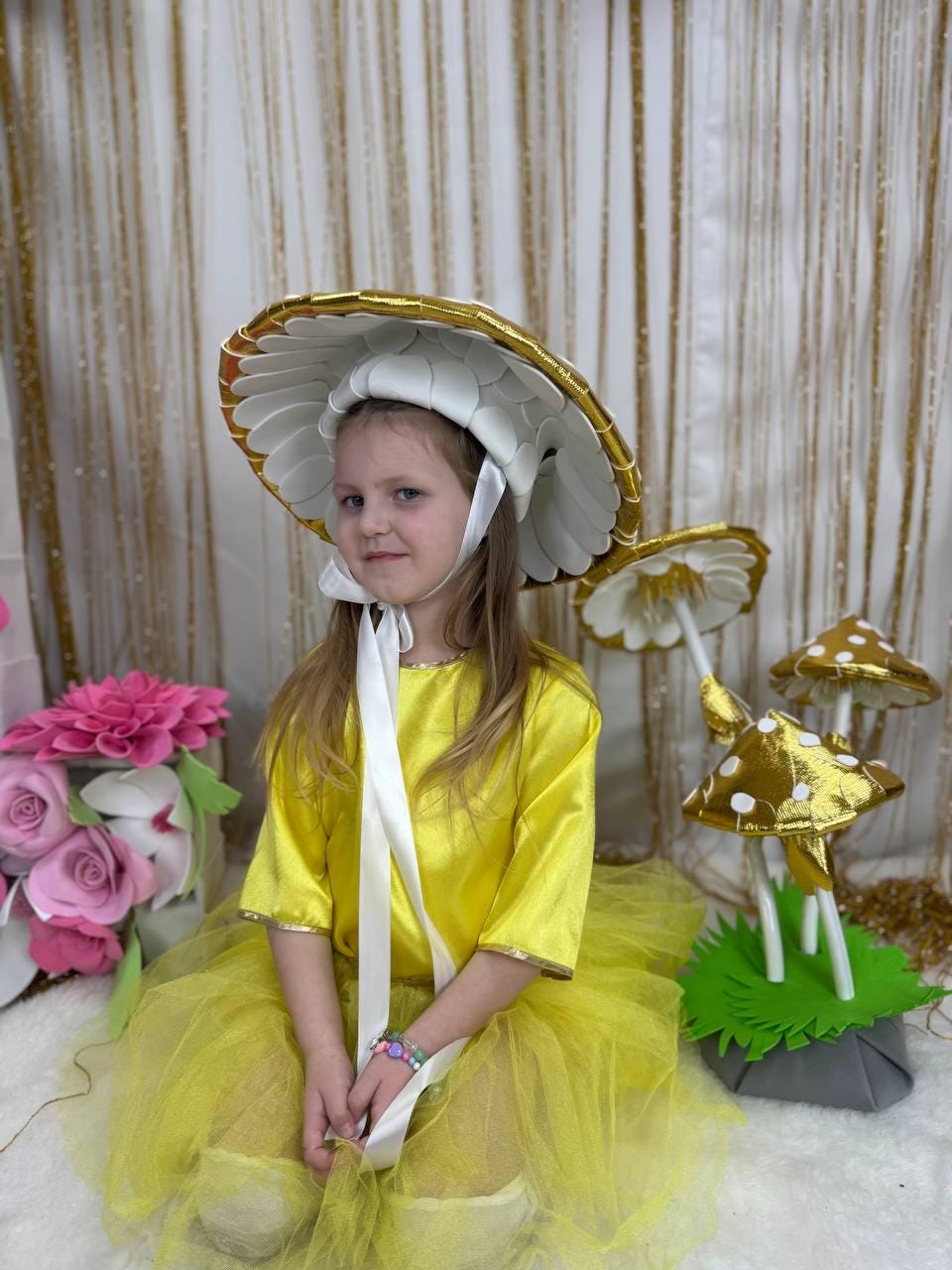 May include: A young girl wearing a yellow dress and a white mushroom hat with gold trim. She is sitting on a white surface with a green and gray mushroom prop behind her.