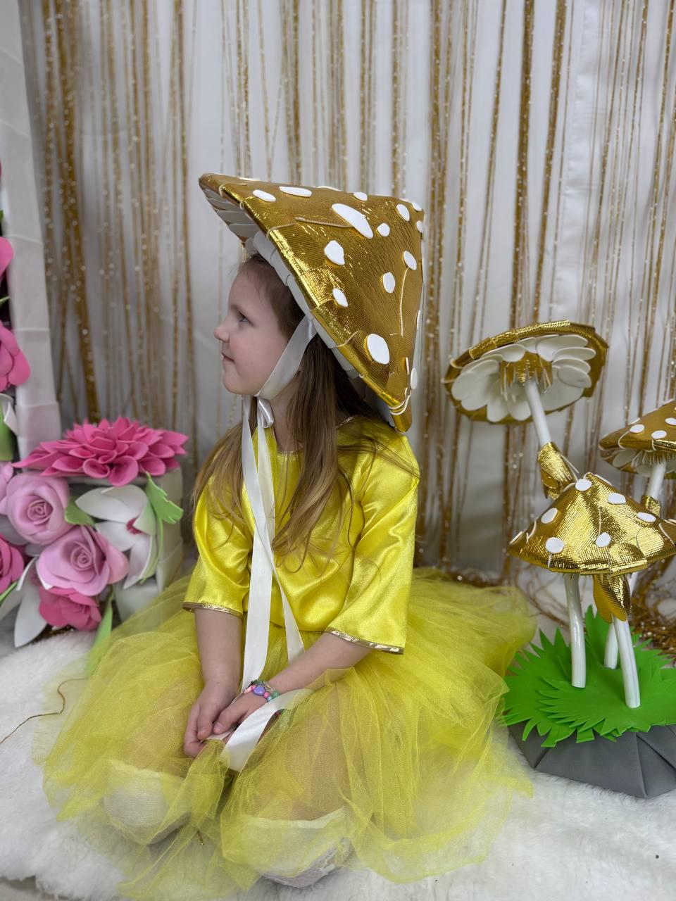 May include: A young girl wearing a yellow dress and a gold mushroom hat. She is sitting on a white fluffy surface with a green and gray mushroom prop behind her.