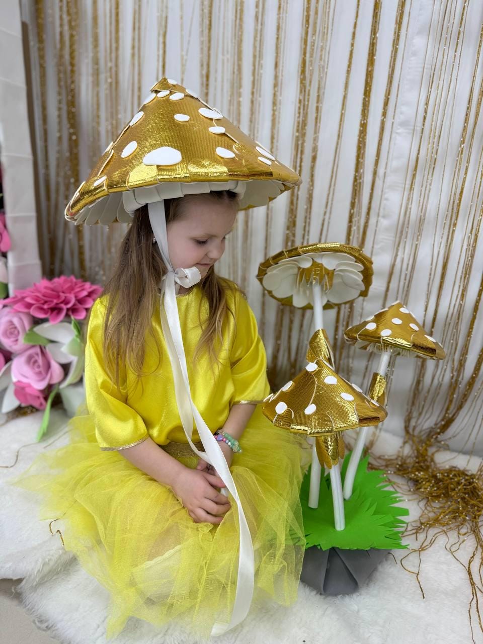 May include: A young girl wearing a yellow dress and a gold mushroom hat sits in front of a set of three gold mushroom decorations. The mushrooms have white polka dots and are on a green base.