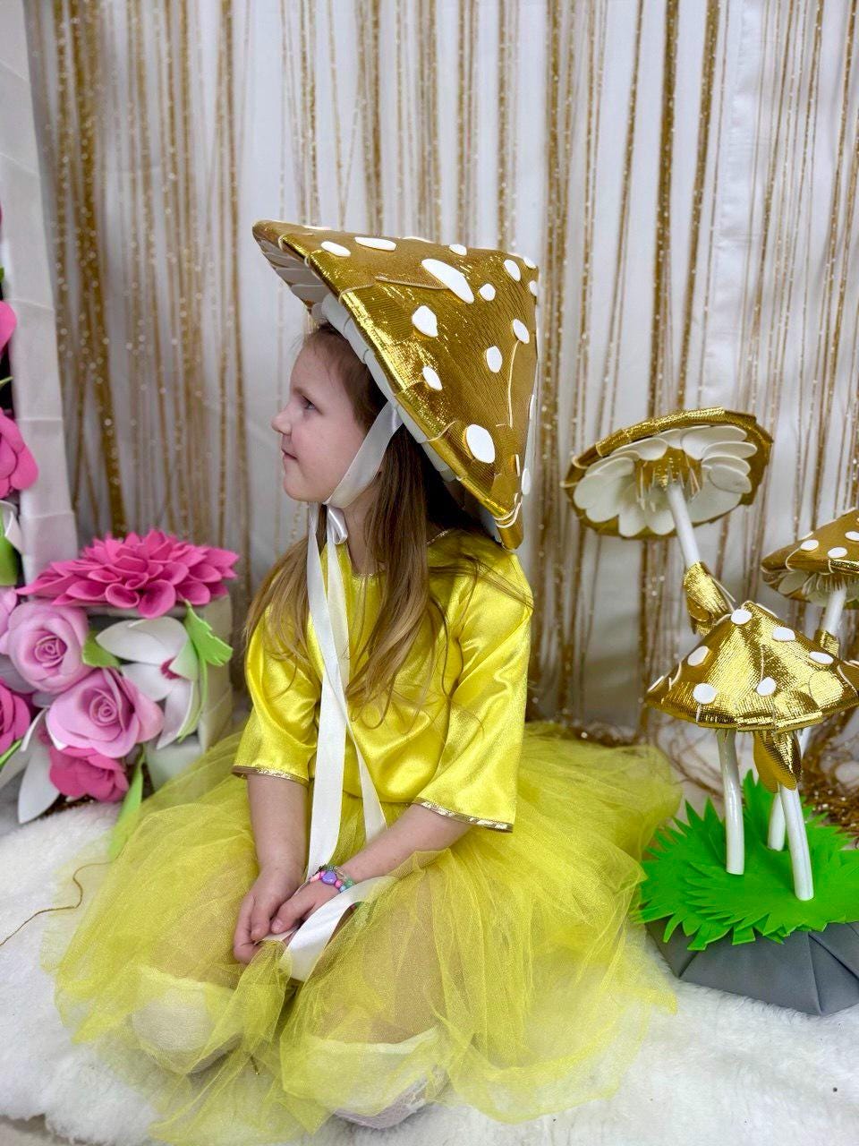 May include: A young girl wearing a yellow dress and a gold mushroom hat. She is sitting on a white fluffy surface with a gold mushroom prop behind her.
