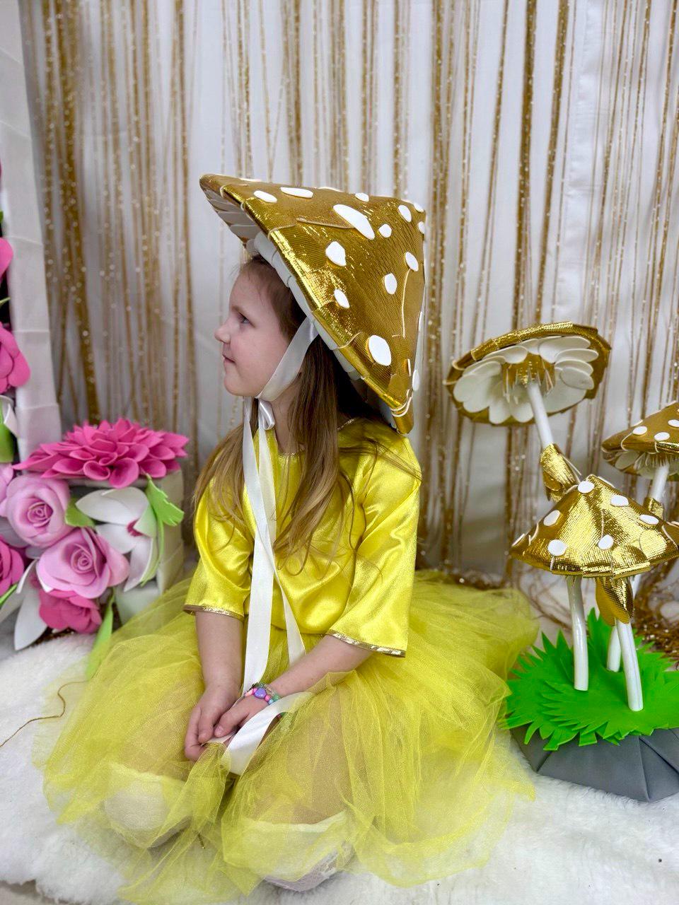 May include: A young girl wearing a yellow dress and a gold mushroom hat. She is sitting on a white fluffy surface with a gold mushroom prop behind her.