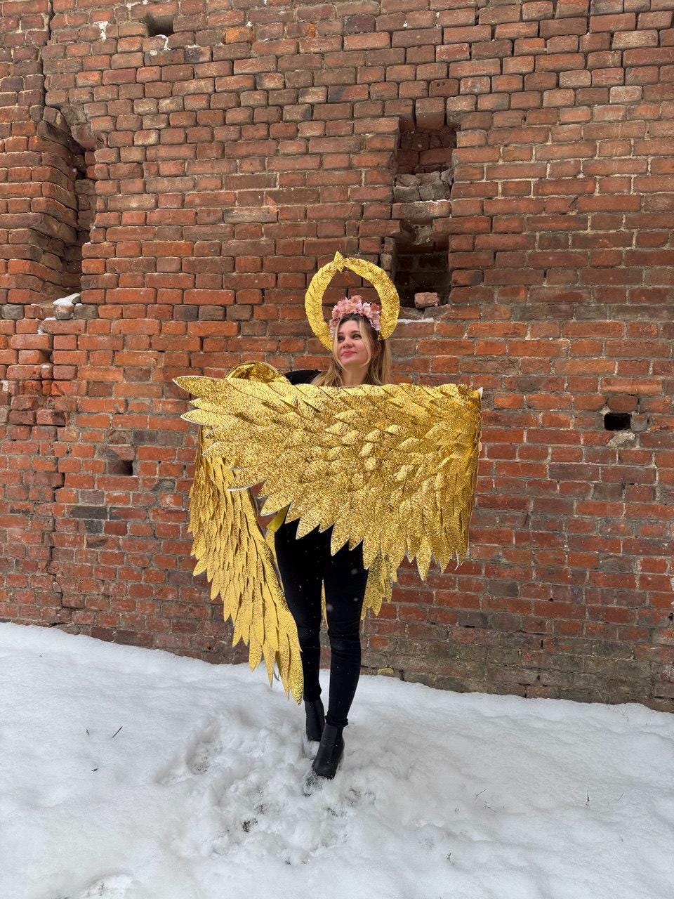 May include: A person wearing a gold angel costume with wings and a halo. The costume is made of feathers and is very detailed. The person is standing in front of a brick wall.