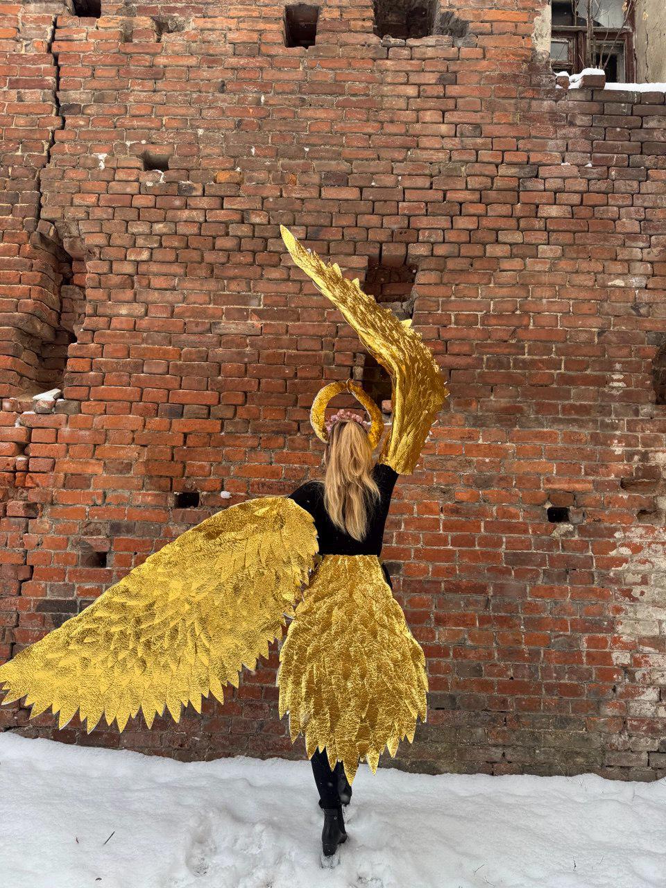 May include: A person wearing a black outfit with large, gold, feathery angel wings. The wings are spread out behind the person, and the person is standing in front of a brick wall.