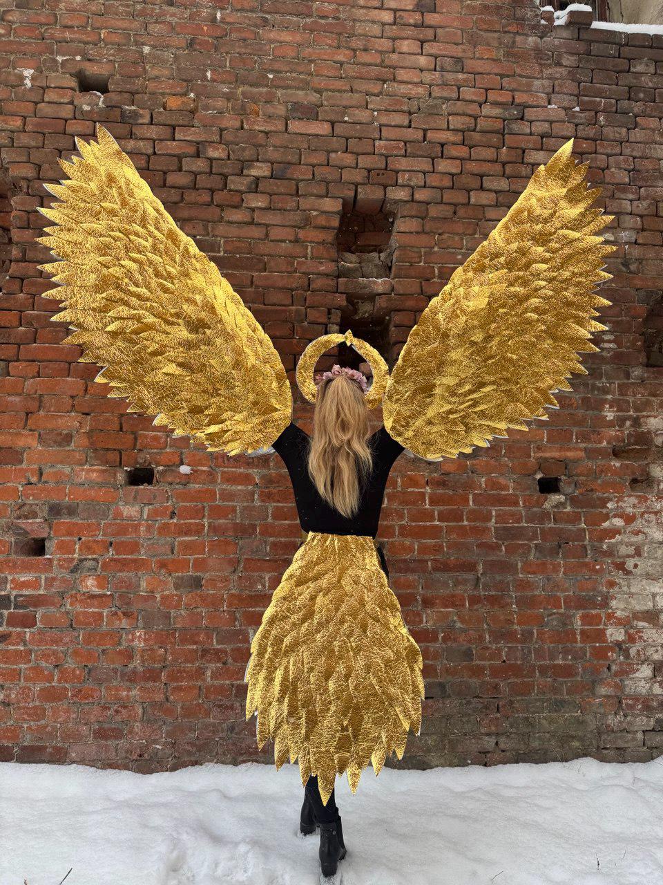May include: A person wearing a black top and a gold angel wing costume with a gold halo. The wings are large and feathery, and the halo is simple and elegant. The person is standing in front of a brick wall.