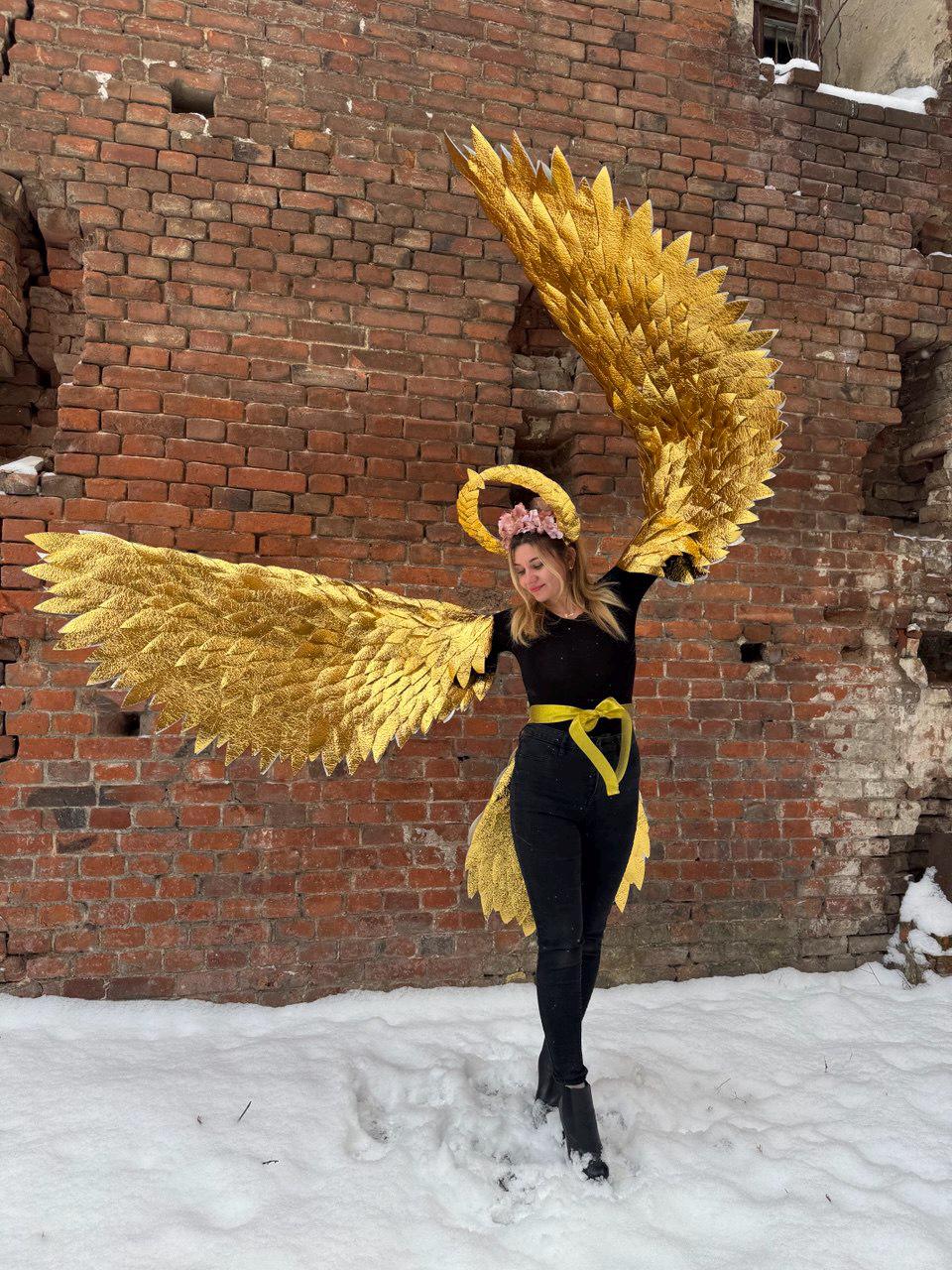 May include: A person wearing a gold angel costume with large, feathery wings. The costume includes a halo and a yellow sash. The person is standing in front of a brick wall with snow on the ground.