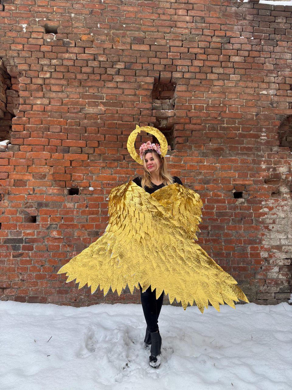 May include: A person wearing a gold angel costume with wings and a flower crown. The costume is made of shiny gold fabric and the wings are large and feathery. The person is standing in front of a brick wall and there is snow on the ground.