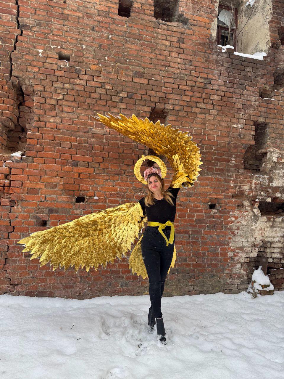 May include: A person wearing a black outfit with a yellow belt and a gold angel wing costume. The wings are large and made of feathers. The person is standing in front of a brick wall with snow on the ground.