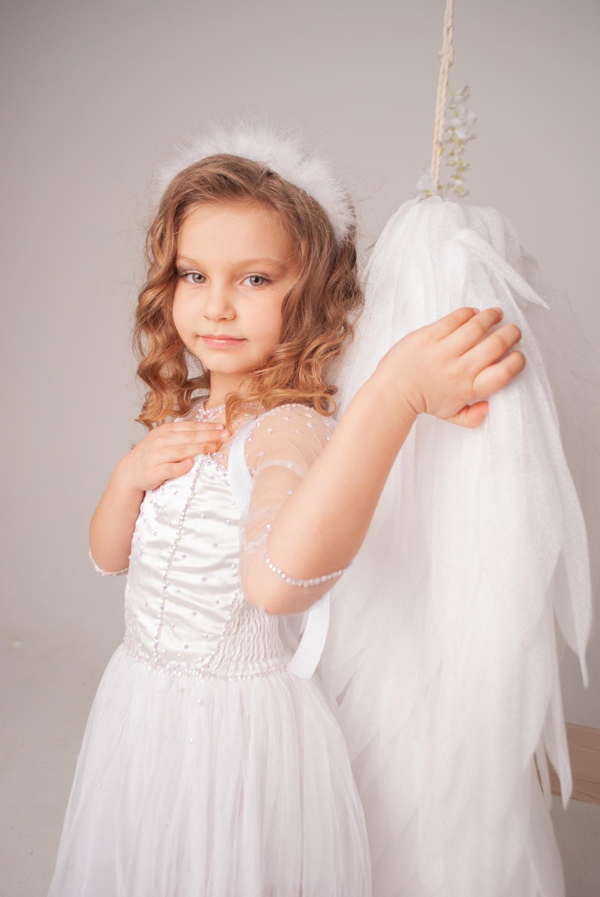 May include: A young girl dressed as an angel wearing a white dress and a white halo. She is holding a white feathery wing.