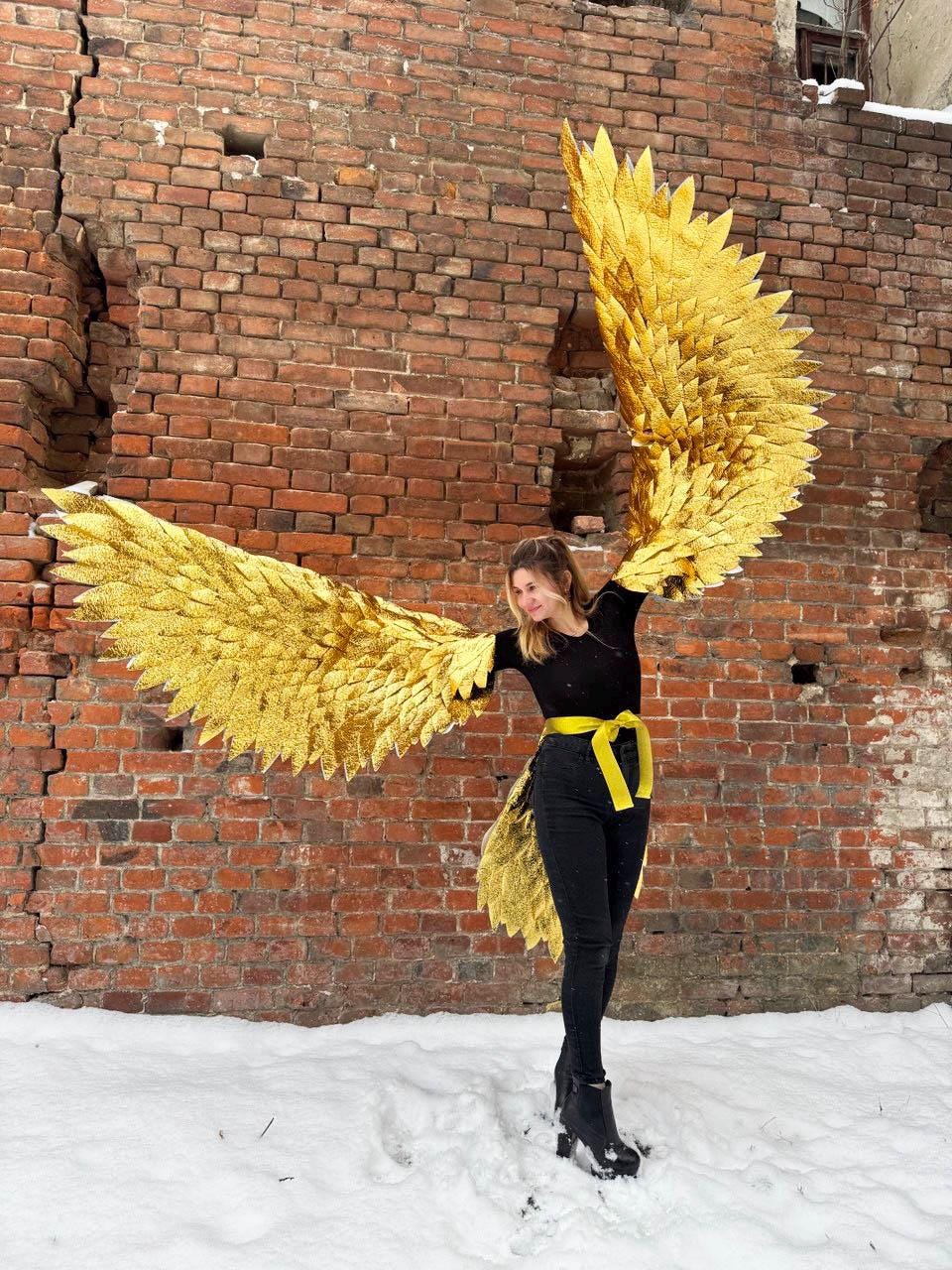 May include: A person wearing a black top and black pants with a yellow belt, holding large gold angel wings in front of a brick wall.