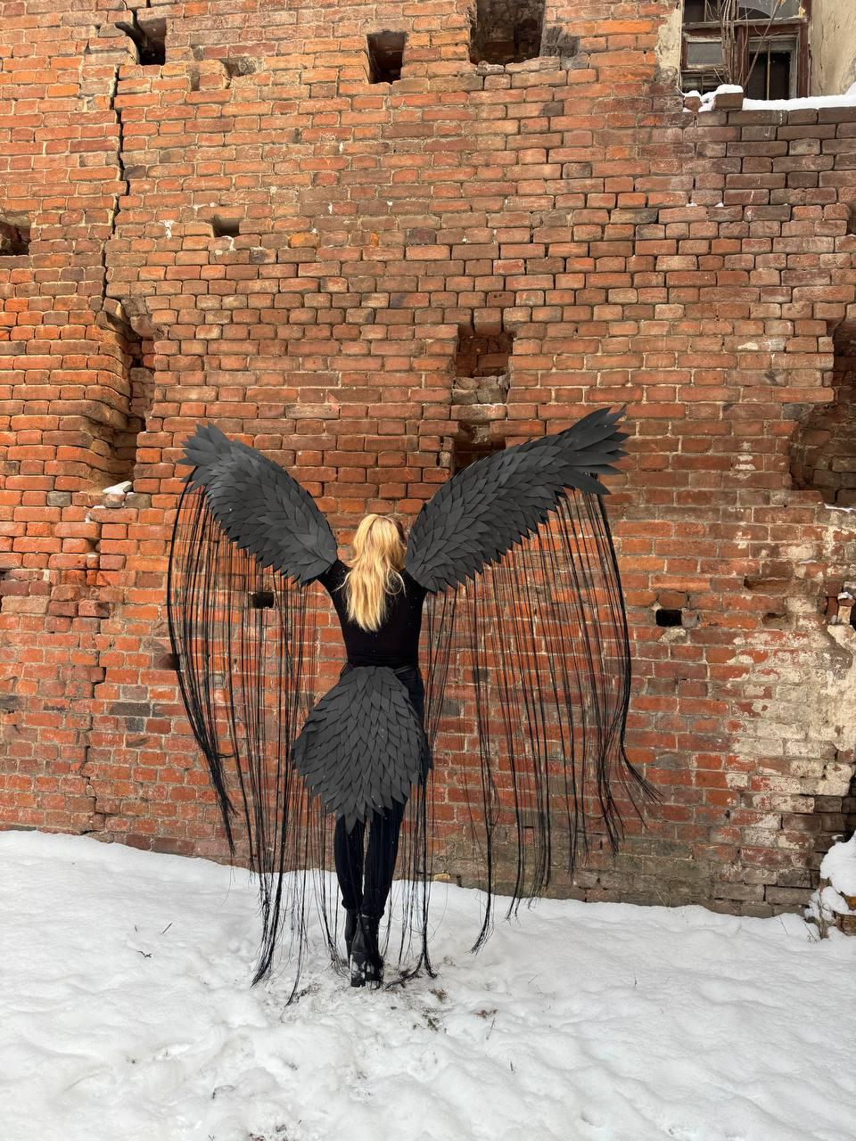 May include: A person wearing a black costume with large, feathery wings. The wings are made of black feathers and are spread out behind the person. The person is standing in front of a brick wall.