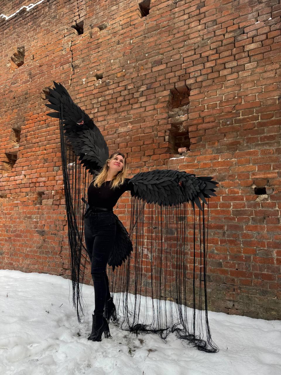 May include: A person wearing a black outfit with large black feathered wings. The wings are attached to the person's back and have long black streamers hanging down. The person is standing in front of a brick wall with snow on the ground.