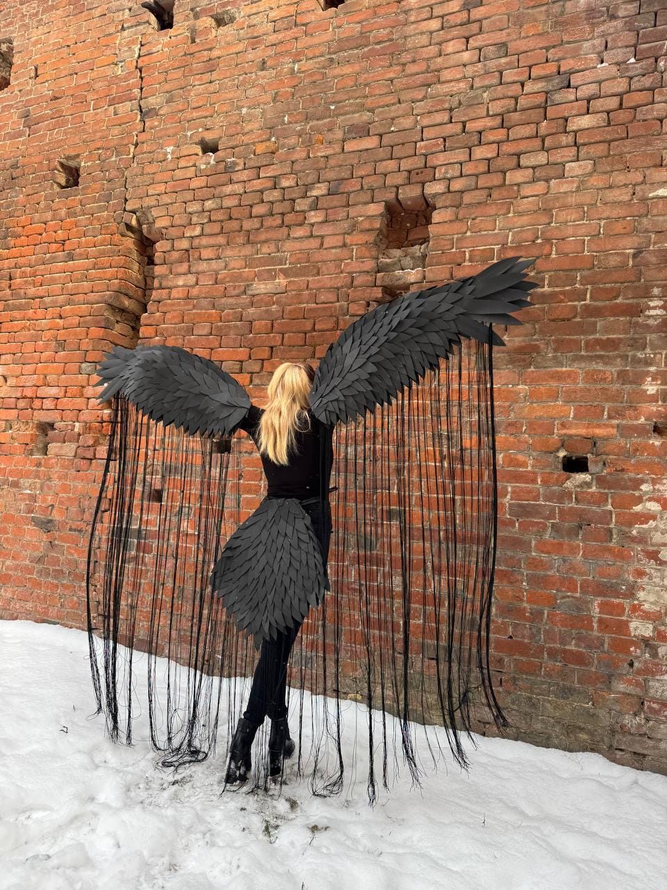May include: A person wearing a black costume with large black wings and a long black tail. The wings are made of feathers and the tail is made of long black strands. The person is standing in front of a brick wall.