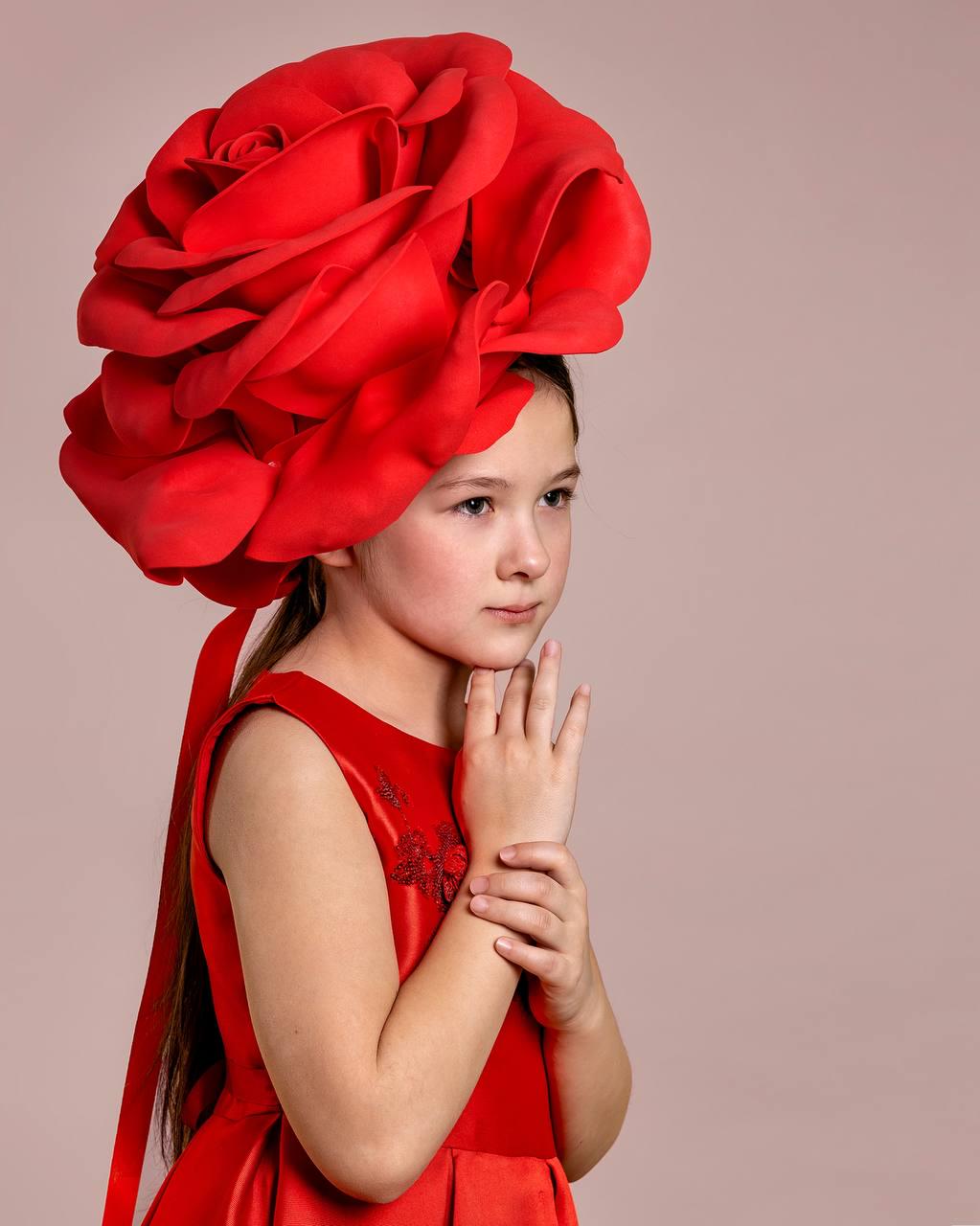 May include: A young person wearing a vibrant red dress and a large, elaborate red rose headpiece. The headpiece is the focal point, with multiple layers of fabric creating the rose petals. The dress has a sleeveless design and a subtle floral embroidery.