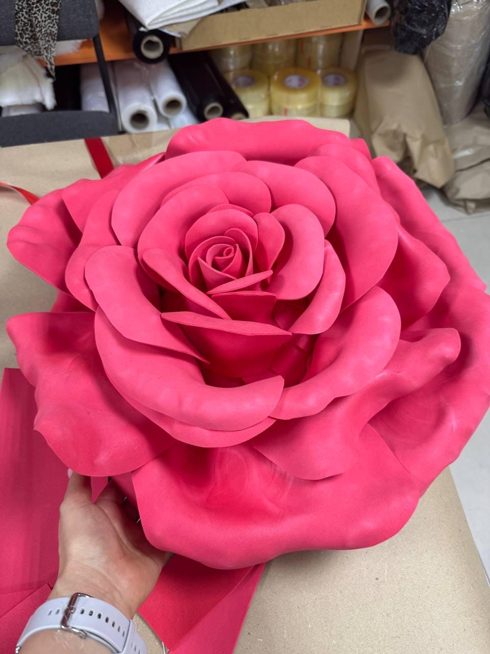 May include: A large, vibrant pink rose made of fabric. The petals are layered and detailed, creating a realistic appearance. The rose is held by a hand, with a pink ribbon attached. The background is a workshop setting.