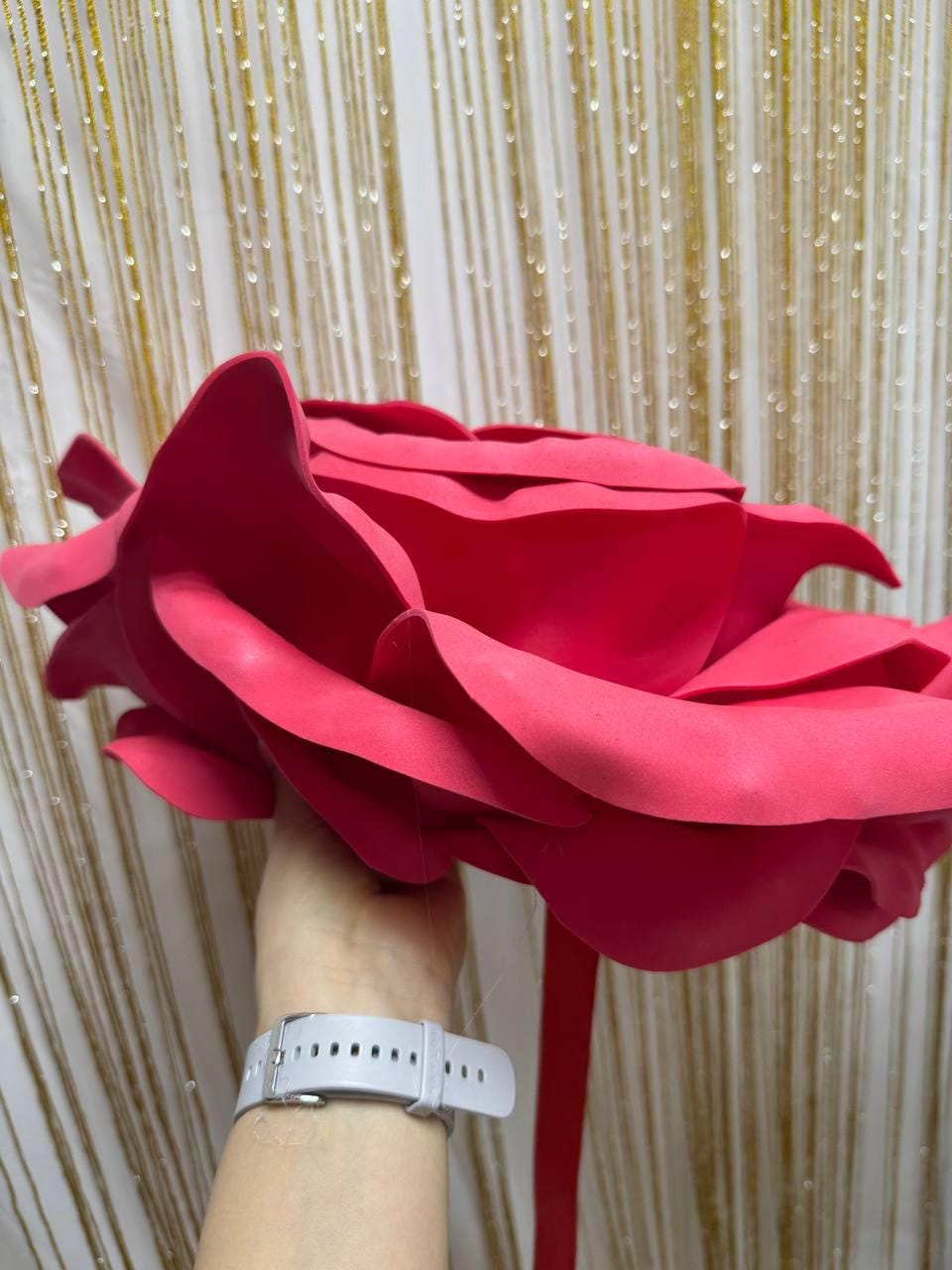 May include: A large, vibrant red paper rose with multiple layers of petals. The oversized flower is held by a person, showcasing its size and detail. The background features gold and white vertical lines.