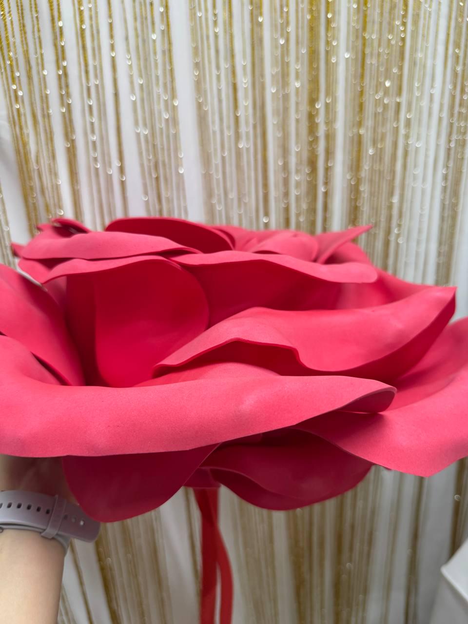 May include: A large, vibrant red paper flower with layered petals. The flower is held by a person, with a gold and white background. The flower's petals are a deep shade of red, creating a striking visual.