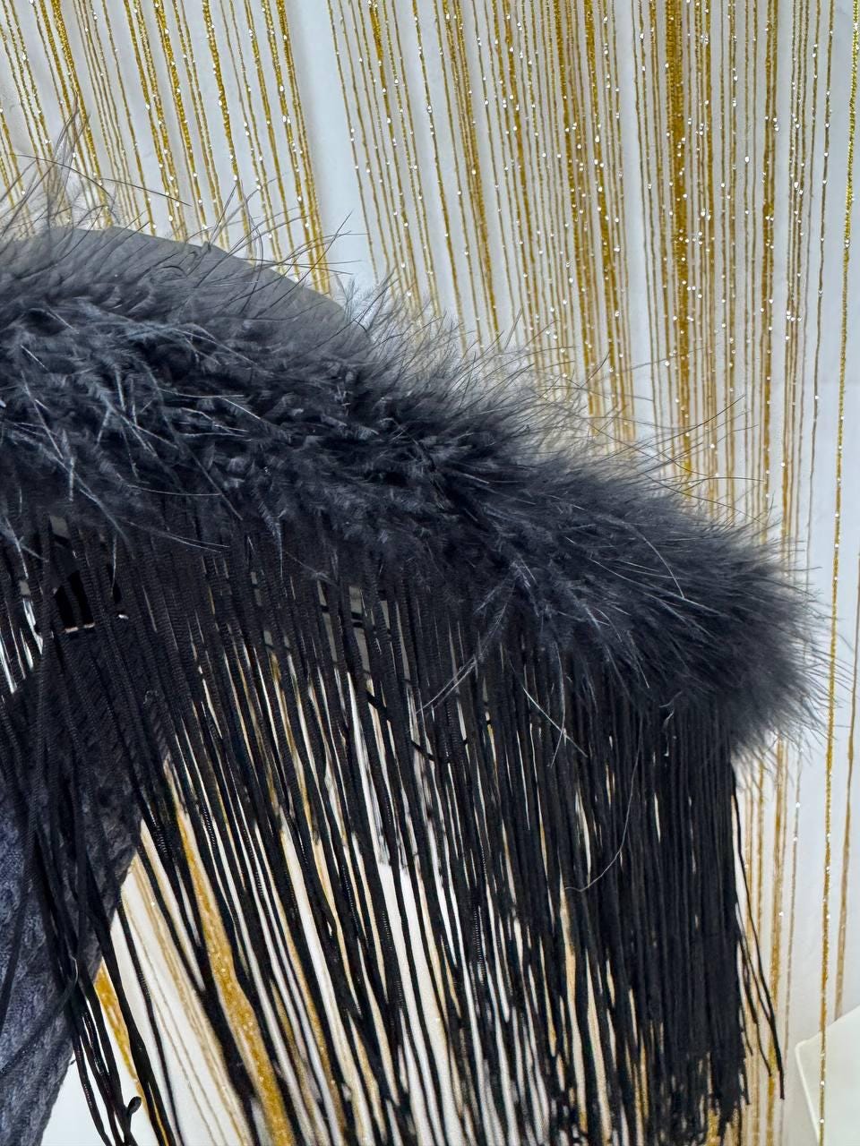 May include: A dark gray hat with a fluffy feather trim and long black fringe. The hat is set against a gold and white background. The design is elegant and suitable for special occasions.
