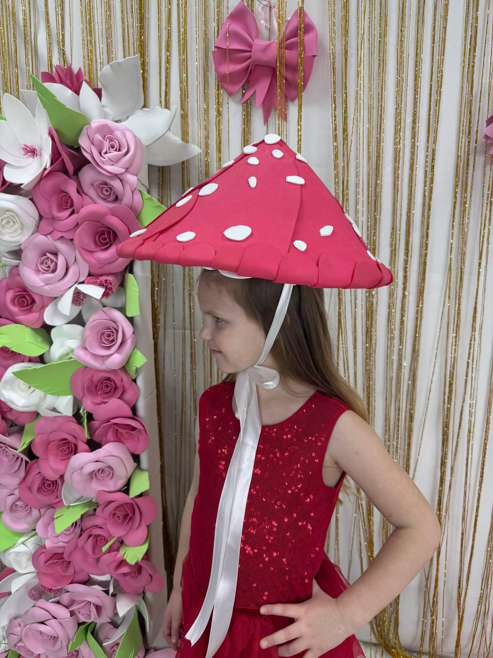 May include: A red mushroom hat with white spots, worn by a person in a red sequined dress. The hat is secured with a white ribbon. A large floral arrangement of pink and white paper flowers is in the background.