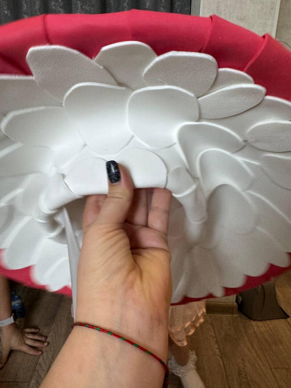 May include: A large, mushroom-shaped prop with a red top and white, petal-like structures underneath. The prop is held by a hand with a black glitter nail. The overall design suggests a costume or decorative item.