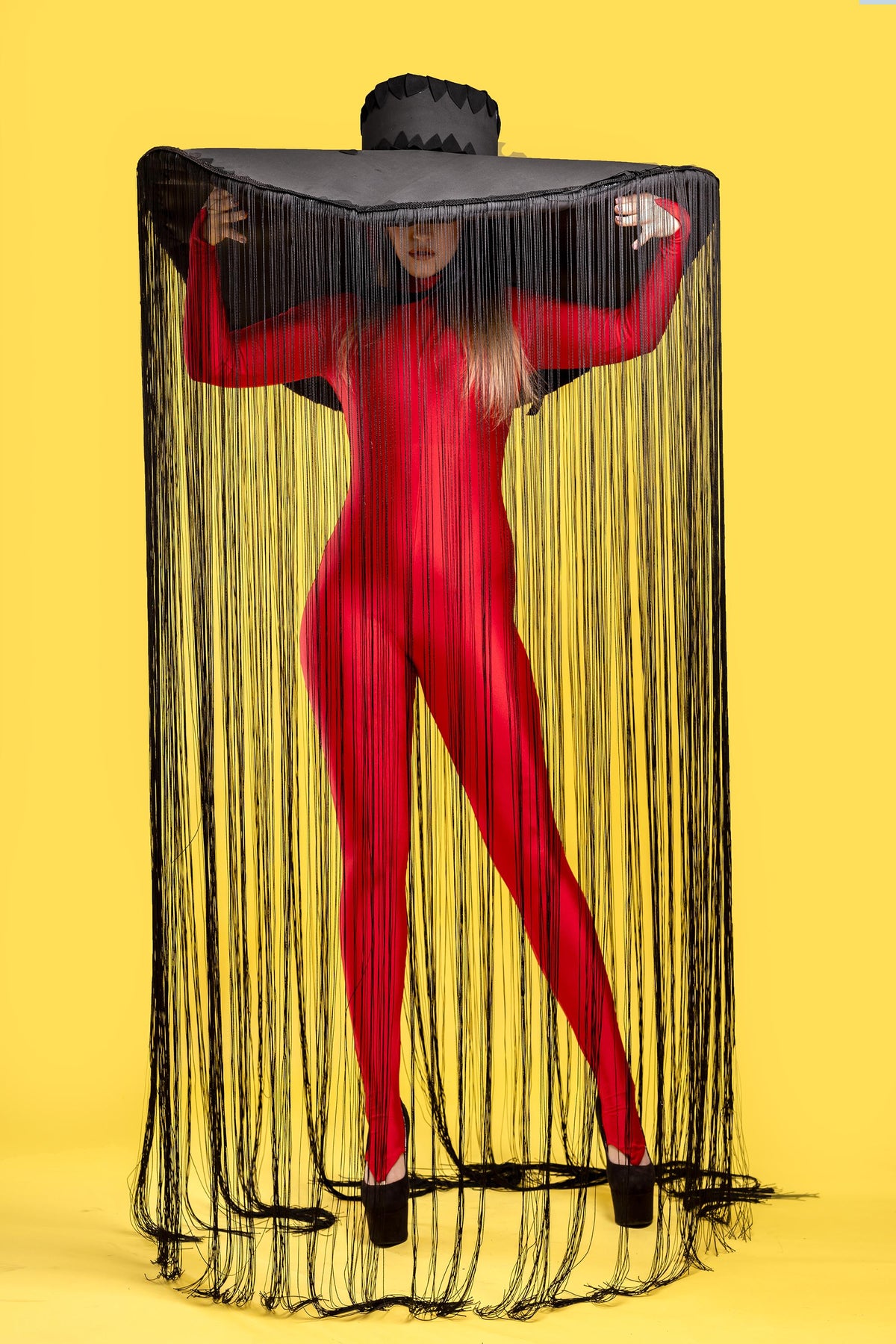 May include: A person wearing a red bodysuit and a large black hat with long black fringe. The hat is covering the person's face. The person is standing on a yellow background.