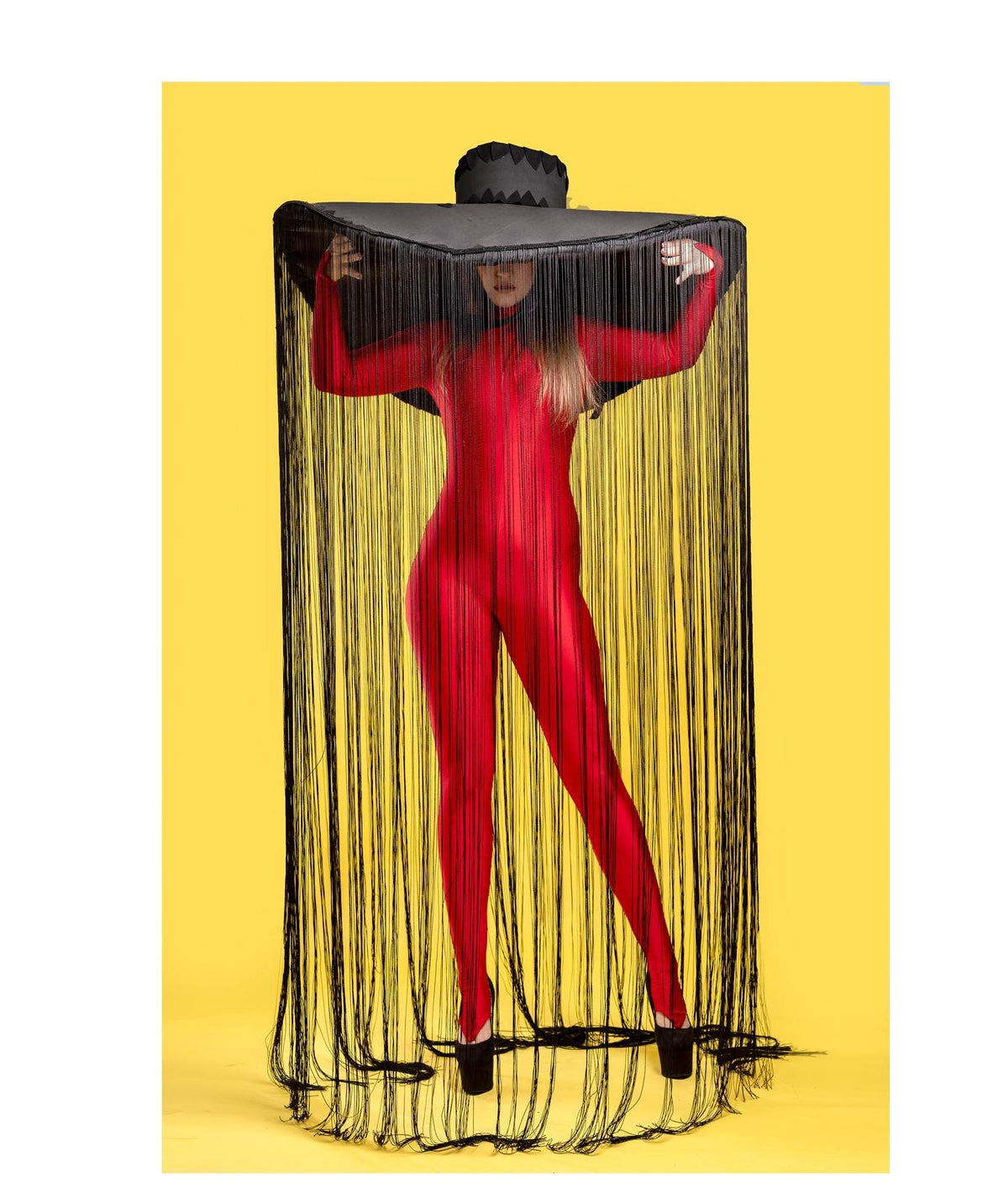 May include: A person wearing a red bodysuit and a large black hat with long fringe. The hat is covering the person's face. The background is yellow.