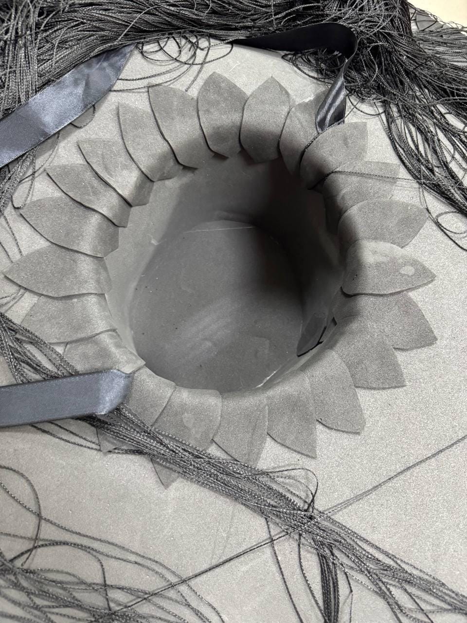 May include: A black hat with a brim made of black fringe. The crown of the hat is made of black fabric with a circular opening in the center. The opening is surrounded by a ring of black fabric petals.