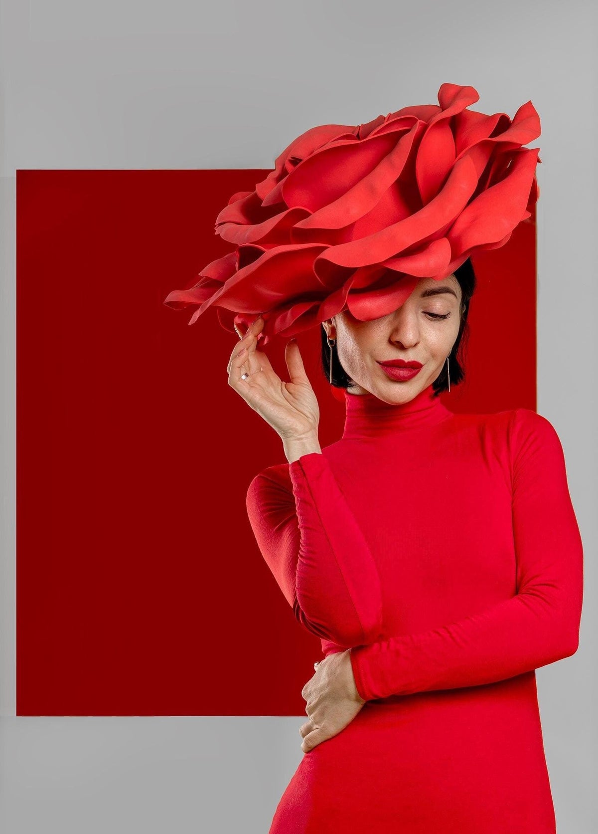 May include: A woman wearing a red dress and a large, red, rose-shaped hat. The hat is covering most of her face. She is standing in front of a red background.
