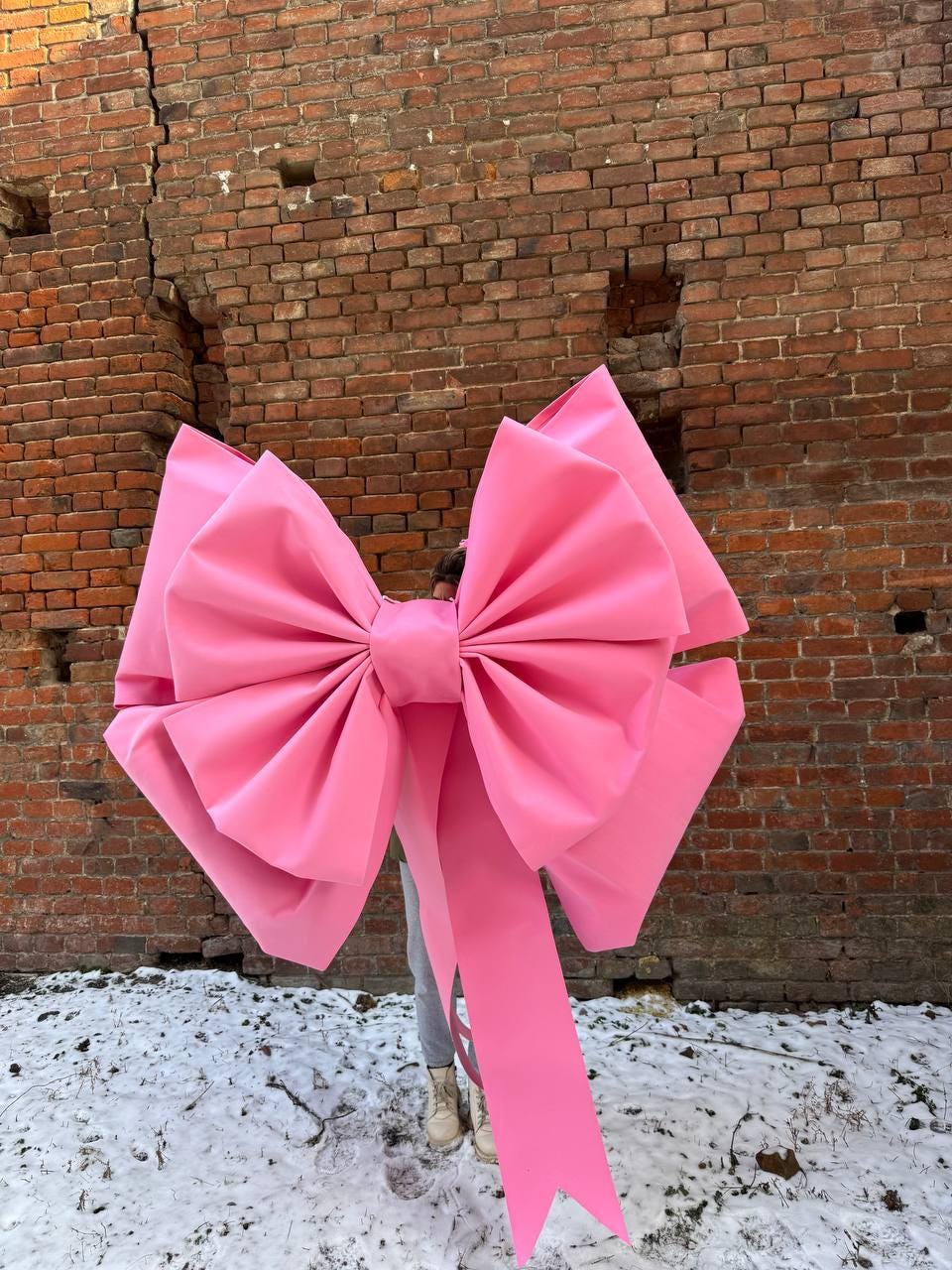 May include: A large pink bow with a long tail. The bow is made of fabric and is tied in a knot. The bow is in front of a brick wall.