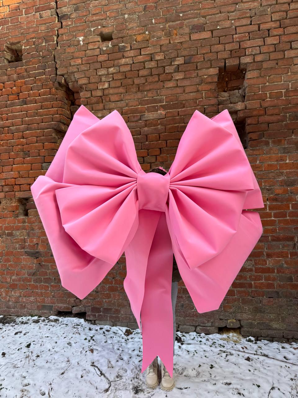 May include: A large pink bow made of fabric, with a long tail, is positioned in front of a brick wall. The bow is so large that it covers the person standing behind it.
